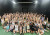 The cast of the 2023 musical gathered for a group shot, looking happy with their hands in the air in the Drama Centre during a rehearsal